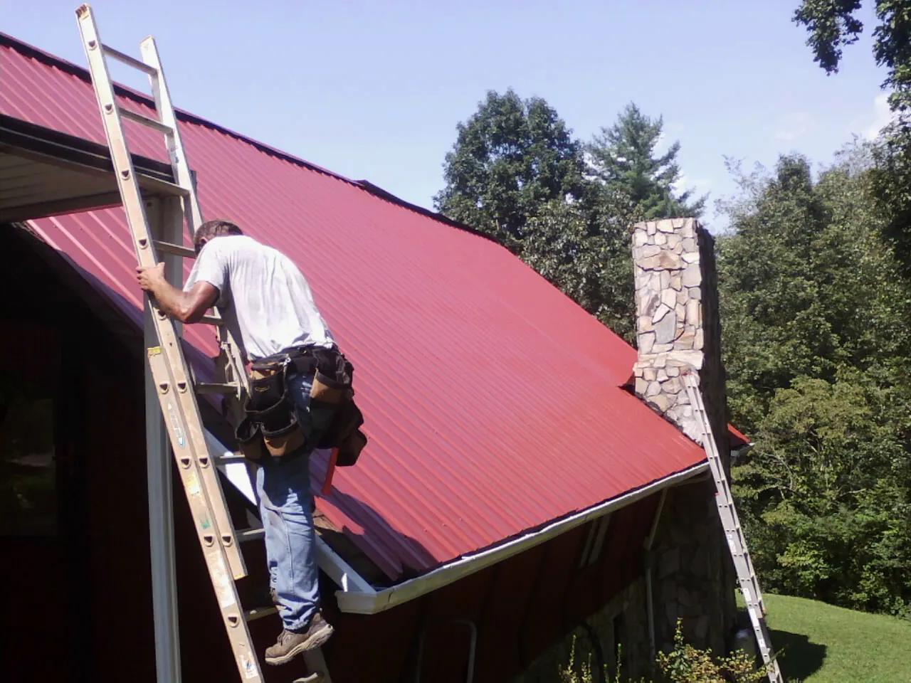 Roofer on a ladder beside a red metal roof and stone chimney — Soffit & Fascia Repair by Roofing Brothers in Maybrook